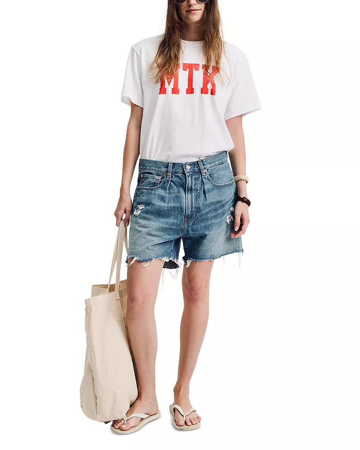 A person holds a beige tote bag while wearing a white T-shirt with red letters, Denimist's BROOKE SHORT denim shorts featuring frayed hems and a five-pocket style, and white flip-flops. They have long hair and wear a wristwatch.