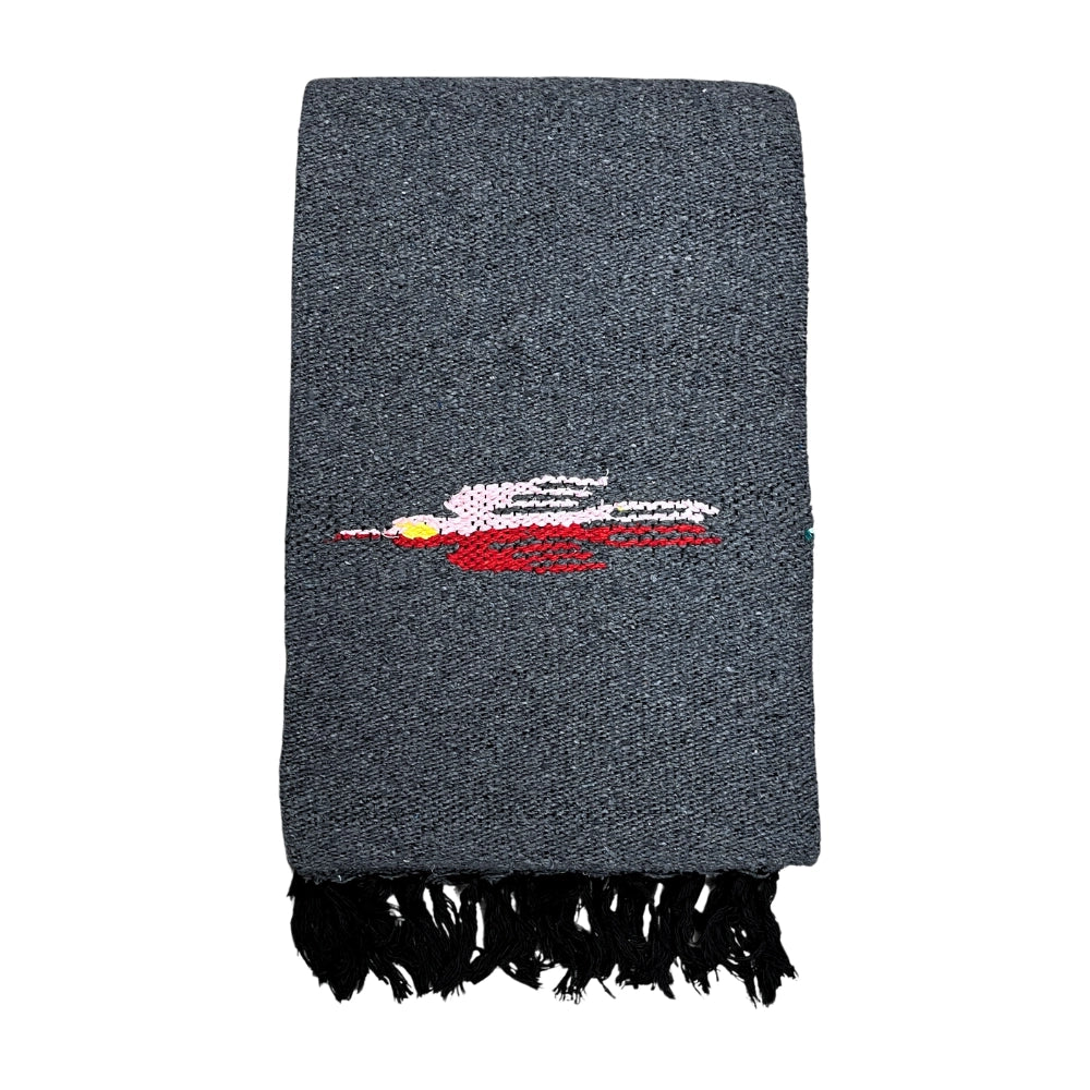 The Faire Charcoal Thunderbird Baja Blanket is a folded, dark gray woven scarf with black fringe and an embroidered pink, white, and red figure—evoking the handmade style of traditional Mexican Thunderbird blankets.