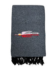 The Faire Charcoal Thunderbird Baja Blanket is a folded, dark gray woven scarf with black fringe and an embroidered pink, white, and red figure—evoking the handmade style of traditional Mexican Thunderbird blankets.