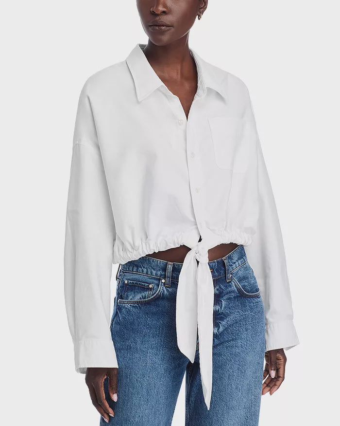 A person wearing the Denimist L/S FRONT TIE SHIRT WHITE S26, a 100% cotton, long-sleeved button-up with a point collar tied at the waist, and blue jeans, stands against a plain white background.