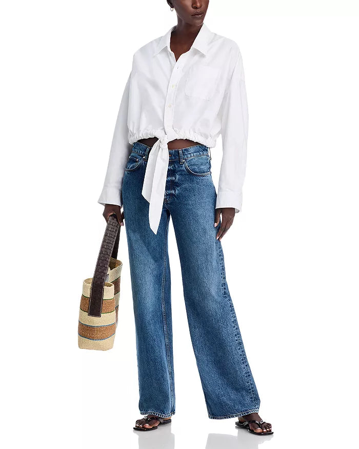 A person wears the Denimist L/S FRONT TIE SHIRT WHITE S26, tied at the waist, with blue wide-leg jeans, black sandals, and a woven striped tote bag, standing against a white background.