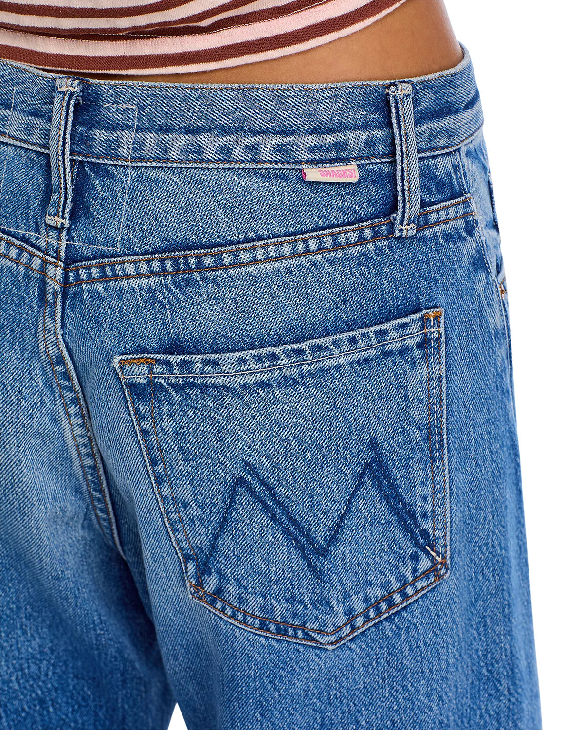 Close-up of the back of a person wearing Mother’s The Lemon Twist Sneak blue denim high rise jeans with a large “M” stitched on the five-pocket style, and a striped shirt partially visible at the waist.