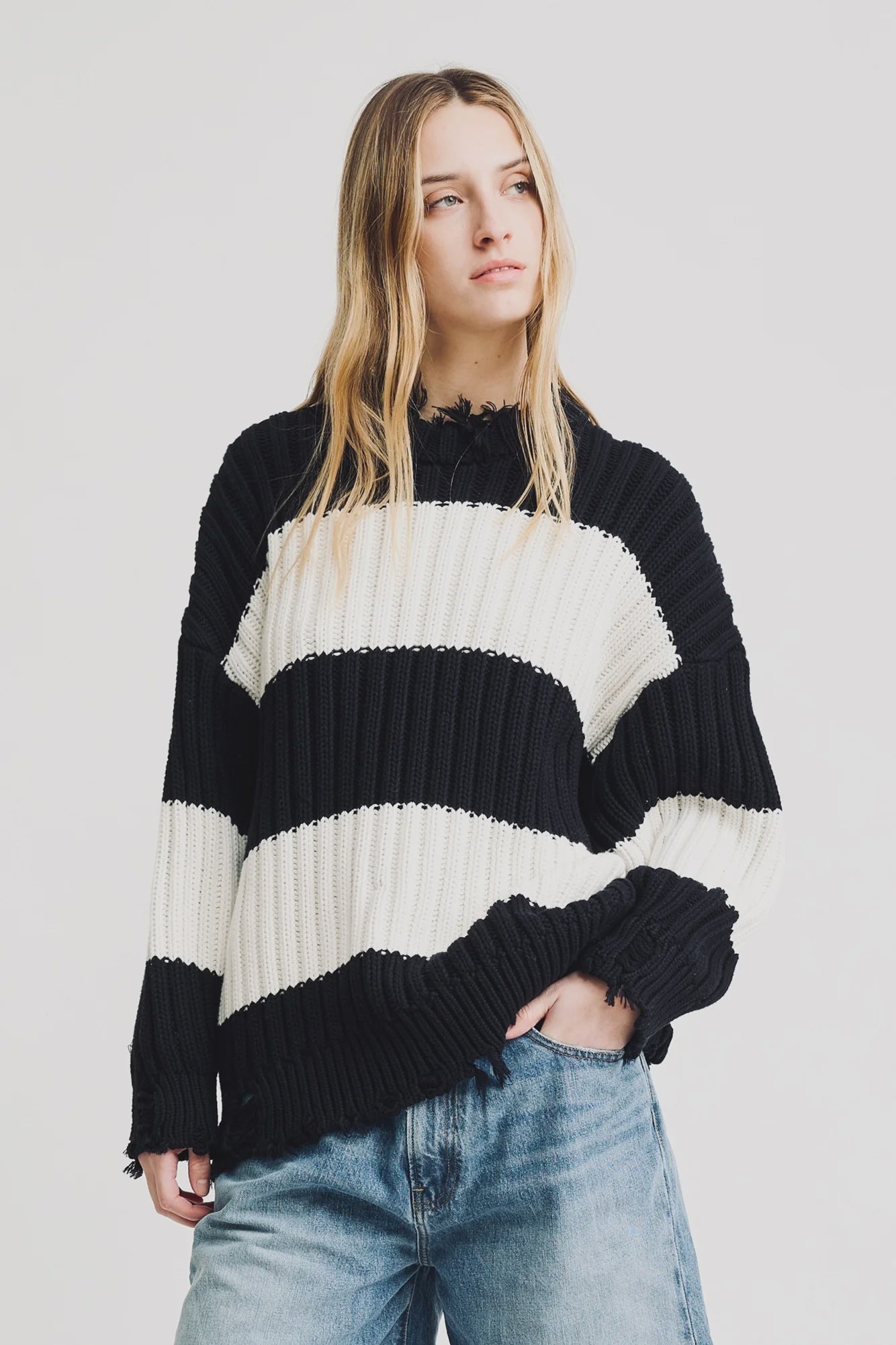 A young woman with long blonde hair wears the Denimist OVERSIZED RIBBED SWEATER in black and white stripes, paired with blue jeans. She stands against a plain white background, looking slightly to the side with a neutral expression.