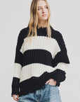 A young woman with long blonde hair wears the Denimist OVERSIZED RIBBED SWEATER in black and white stripes, paired with blue jeans. She stands against a plain white background, looking slightly to the side with a neutral expression.
