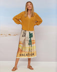 A woman smiles by the beach, wearing the Me369 Elizabeth Midi Skirt—a pleated design with palm tree and "VACATION" print—paired with a mustard cardigan and tan sandals for a perfect vintage-inspired summer look.
