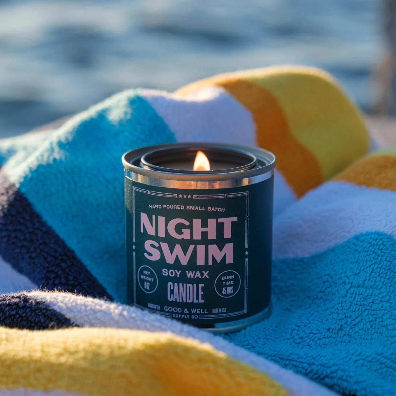 A lit G&W Soy Candle by Faire from the Happy Place Collection rests on a colorful striped towel, with a blurred watery backdrop that evokes nostalgic joy and refreshing fragrance.