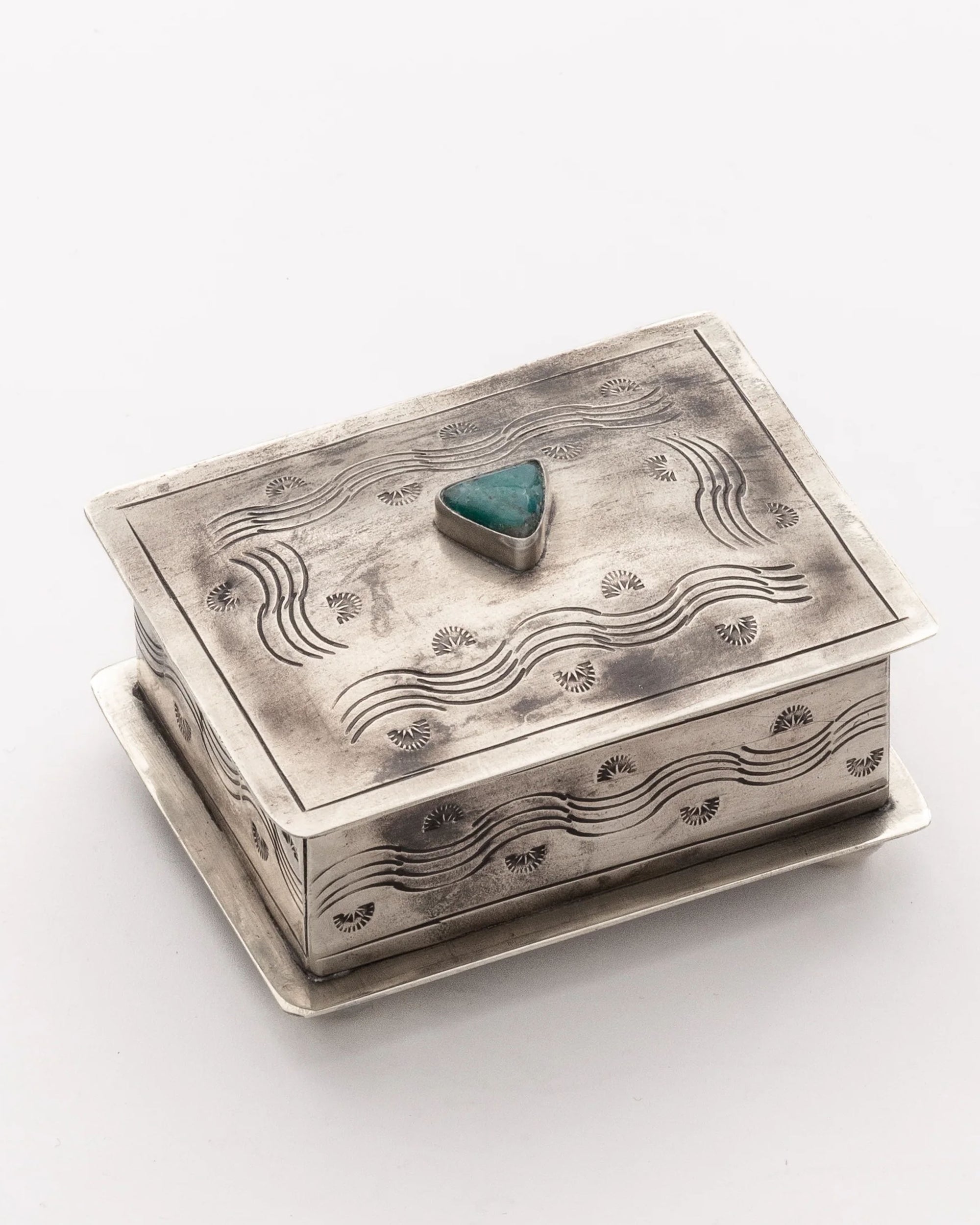 The J Alexander Rustic Silver Small Stamped Box w/ Turquoise features engraved wave and fan motifs with a raised turquoise stone at the center of the lid, resting on a flat silver base against a white background.