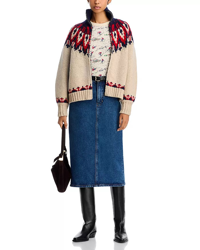 A person wears The Nordic Sweater by The Great Inc., featuring cream with red and navy Fair Isle patterns, paired with a printed shirt, blue denim midi skirt, black knee-high boots, and a dark handbag.