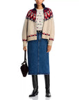 A person wears The Nordic Sweater by The Great Inc., featuring cream with red and navy Fair Isle patterns, paired with a printed shirt, blue denim midi skirt, black knee-high boots, and a dark handbag.