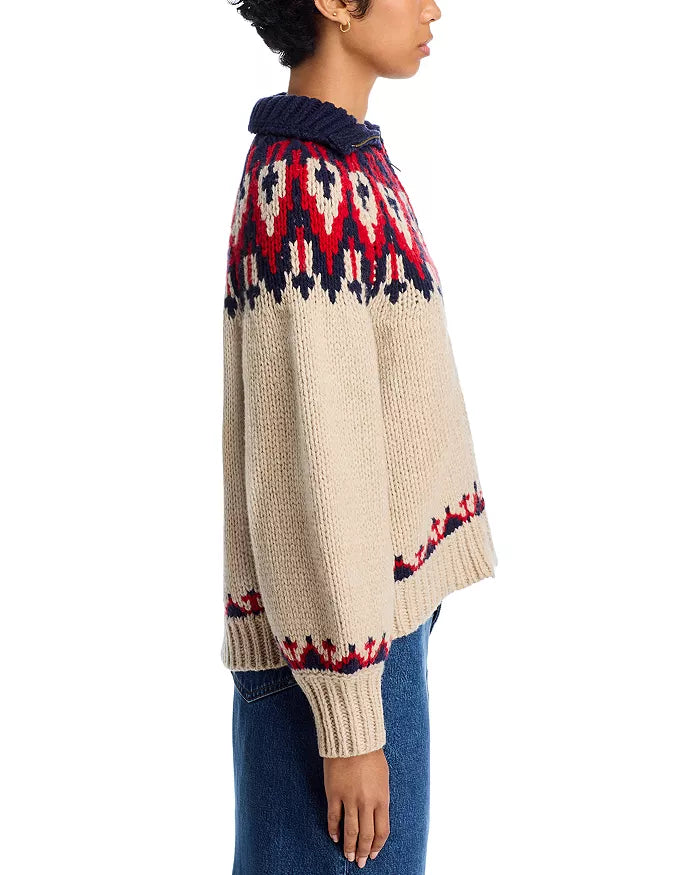 A person stands in profile against a white background wearing The Great Inc.’s The Nordic Sweater, a beige Fair Isle knit featuring red and navy geometric patterns on the yoke, cuffs, and hem. Made from 100% cotton for comfort and warmth.
