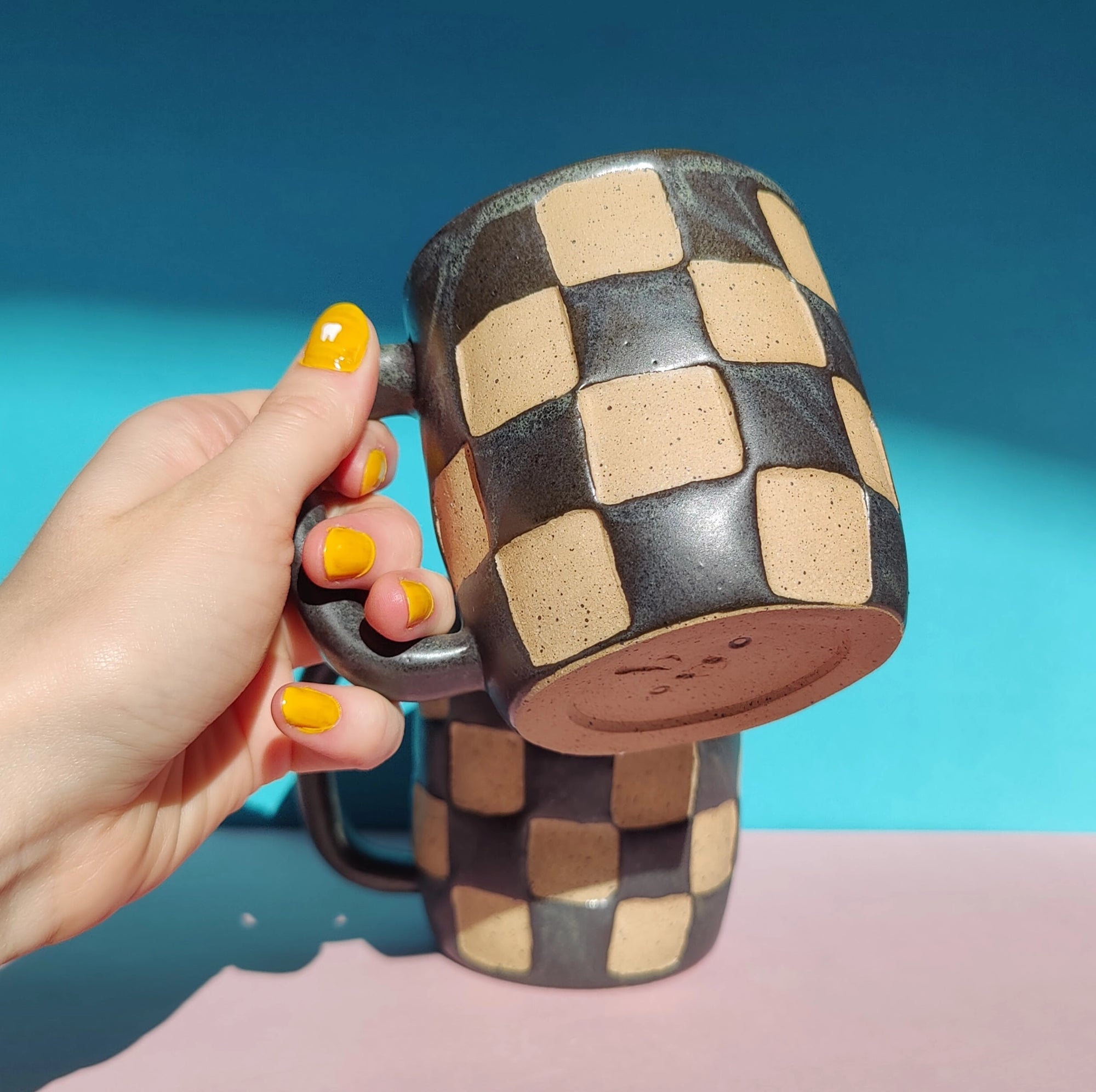 A hand with yellow-painted nails holds the Faire OC Checkerboard Mug, which features a black and beige checkered stoneware design; another matching mug rests on a pink surface against a blue background.