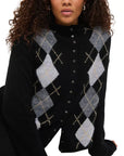 A woman with curly hair wears the Alex Mill Taylor Cardigan Argyle, a black button-up with classic grey and beige argyle on the front. Styled with silver hoop earrings, she is photographed shoulders up, highlighting the cardigan's relaxed fit charm.