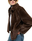 A person wearing the Alex Mill Duane St Jacket in dark brown faux fur, styled with blue jeans, a colorful scarf, and sunglasses, poses with one hand in their pocket against a plain white background.