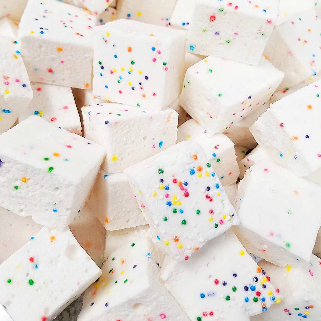 A stack of Faire Funfetti Marshmallows—white, allergy-friendly cubes topped with colorful round sprinkles—are closely arranged together.
