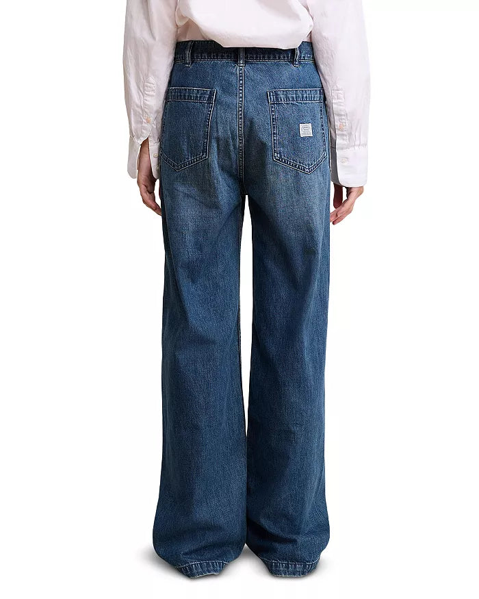 A person stands with their back to the camera, wearing Denimist’s DECK PANT SHANE INDIGO—high rise, wide-leg blue jeans made from 100% cotton with two large back pockets and a small white label on one pocket—paired with a white long-sleeve shirt.