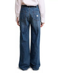 A person stands with their back to the camera, wearing Denimist’s DECK PANT SHANE INDIGO—high rise, wide-leg blue jeans made from 100% cotton with two large back pockets and a small white label on one pocket—paired with a white long-sleeve shirt.