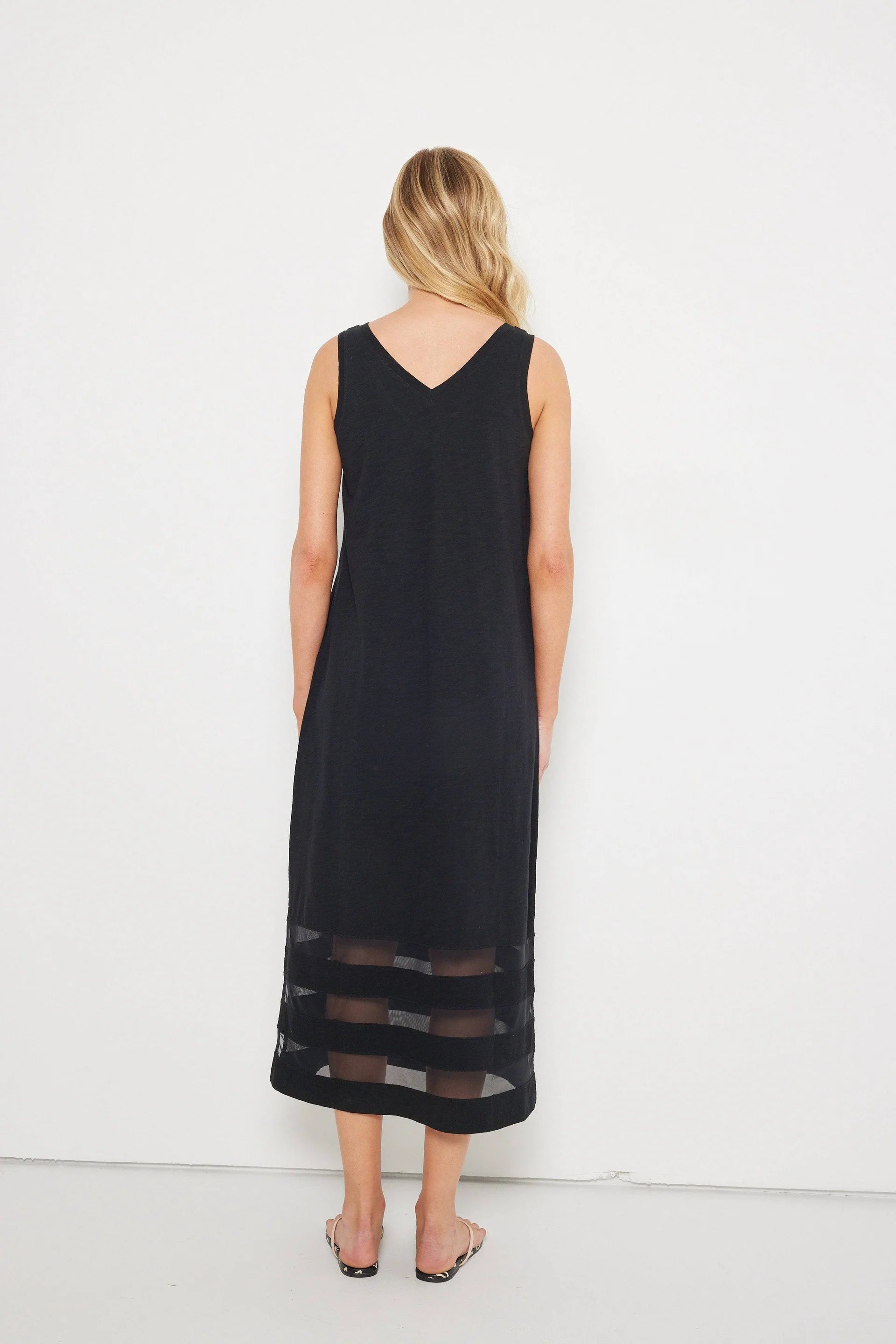 A woman with wavy blonde hair stands barefoot, facing a white wall, wearing the Lisa Todd "Best Dressed" sleeveless black maxi dress with a v-neckline and sheer mesh horizontal stripes at the hem.