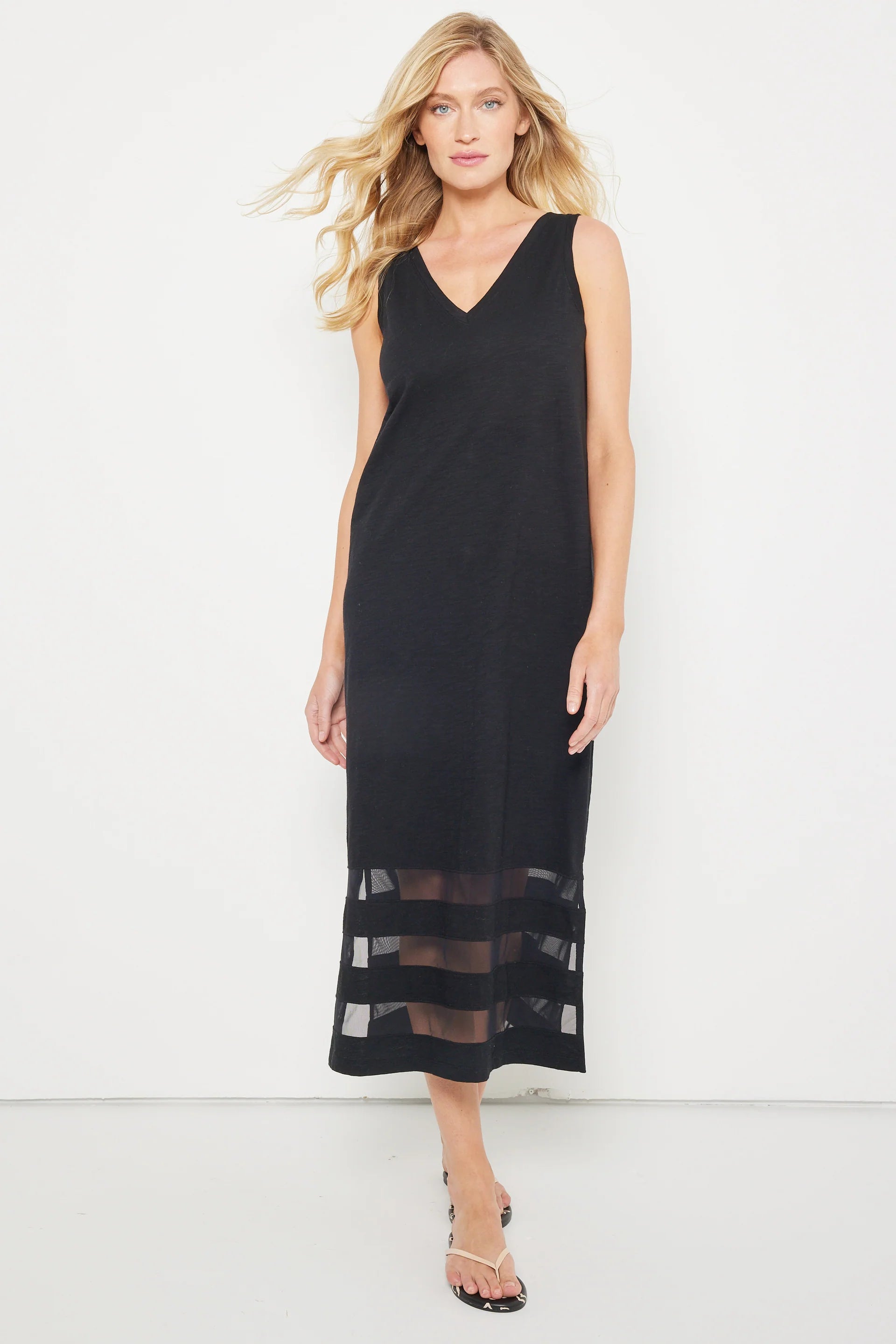 A woman models the "Best Dressed" sleeveless black maxi dress by Lisa Todd, featuring a V-neckline and mesh hem inserts, standing against a white background in sandals and gazing confidently at the camera.