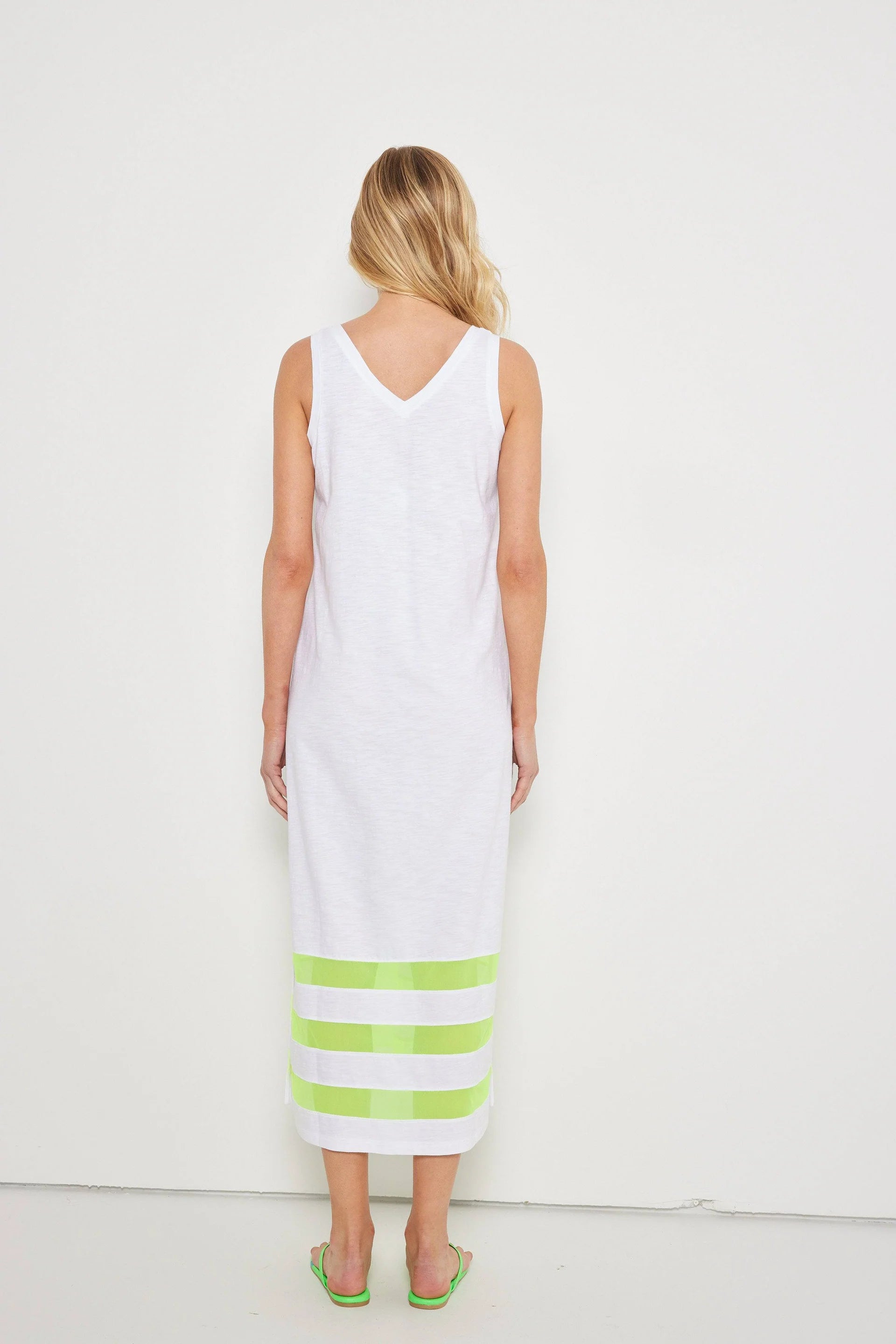 A woman with loose blonde waves faces a white wall, wearing Lisa Todd's "Best Dressed" sleeveless v-neck maxi dress with light green stripes near the hem, paired with matching green sandals.