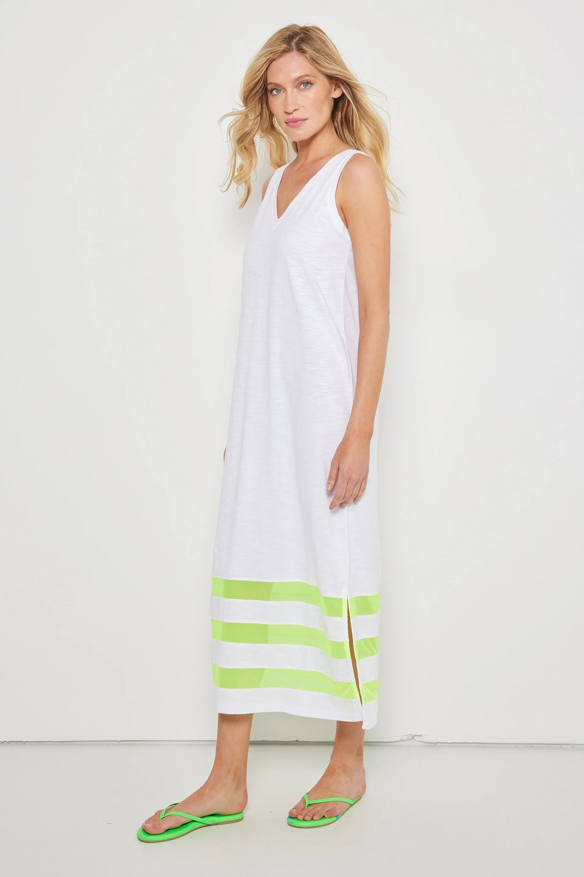 A woman in the Lisa Todd "Best Dressed" sleeveless white maxi dress with a v-neckline and green stripes at the hem, paired with green flip-flops, stands against a plain white background.
