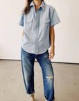 The individual is dressed in The Western Shirt by Hey Gang, a vintage men's light blue short-sleeve button-up with two chest pockets, paired with relaxed-fit jeans featuring a slight rip. The backdrop is an off-white wall and a gray floor, creating a casual and minimalist setting.