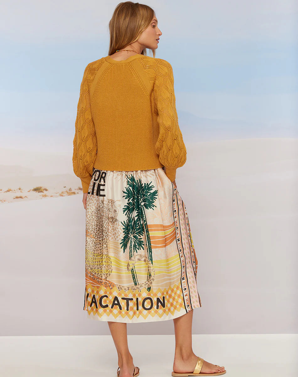A woman faces away in the Me369 Elizabeth Midi Skirt, featuring palm tree and cheetah prints plus "VACATION" at the hem, paired with a mustard sweater and sandals for a chic summer look against a light background.
