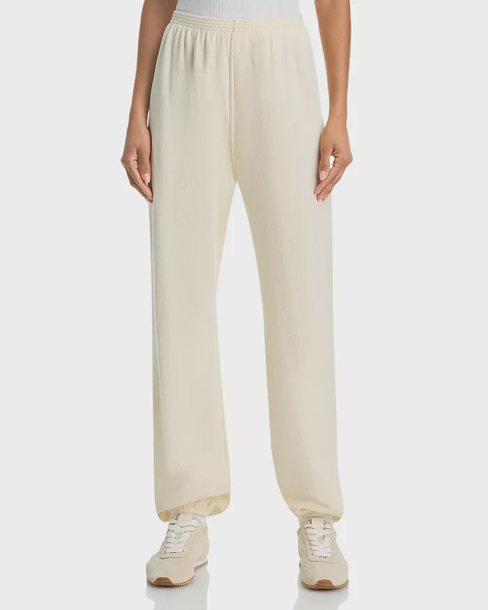 A person wearing The Dance Pant by The Great Inc.—off-white, loose-fitting sweatpants with a straight-leg design—and white sneakers stands against a plain background, shown from waist to feet.