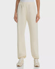 A person wearing The Dance Pant by The Great Inc.—off-white, loose-fitting sweatpants with a straight-leg design—and white sneakers stands against a plain background, shown from waist to feet.
