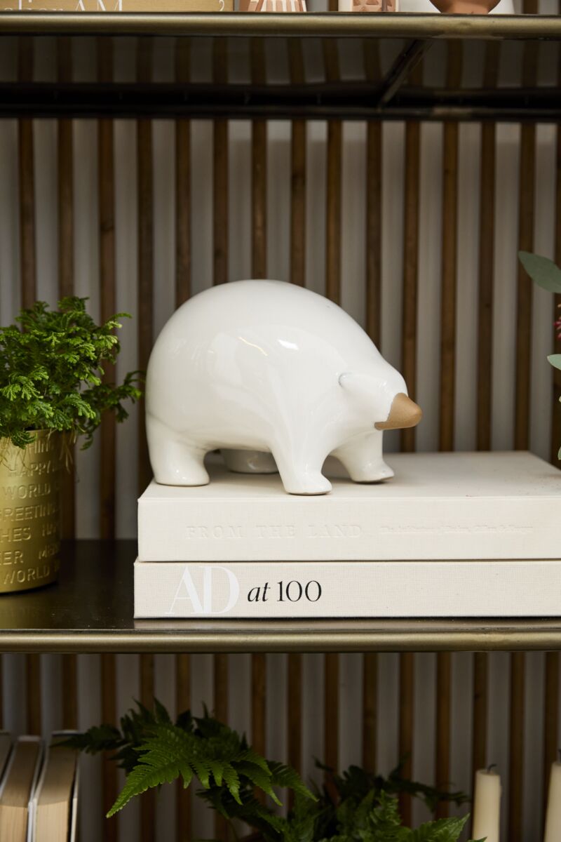 The Accent Decor Ceramic Polar Bear, featuring a brown snout, stands atop a "AD at 100" book and is surrounded by potted plants—a stylish touch for your decorative shelf or Arctic figurine collection.