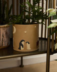 The Accent Decor Antarctica Pot, featuring nativity scene designs, holds a green fern and sits on a dark shelf among other potted plants.