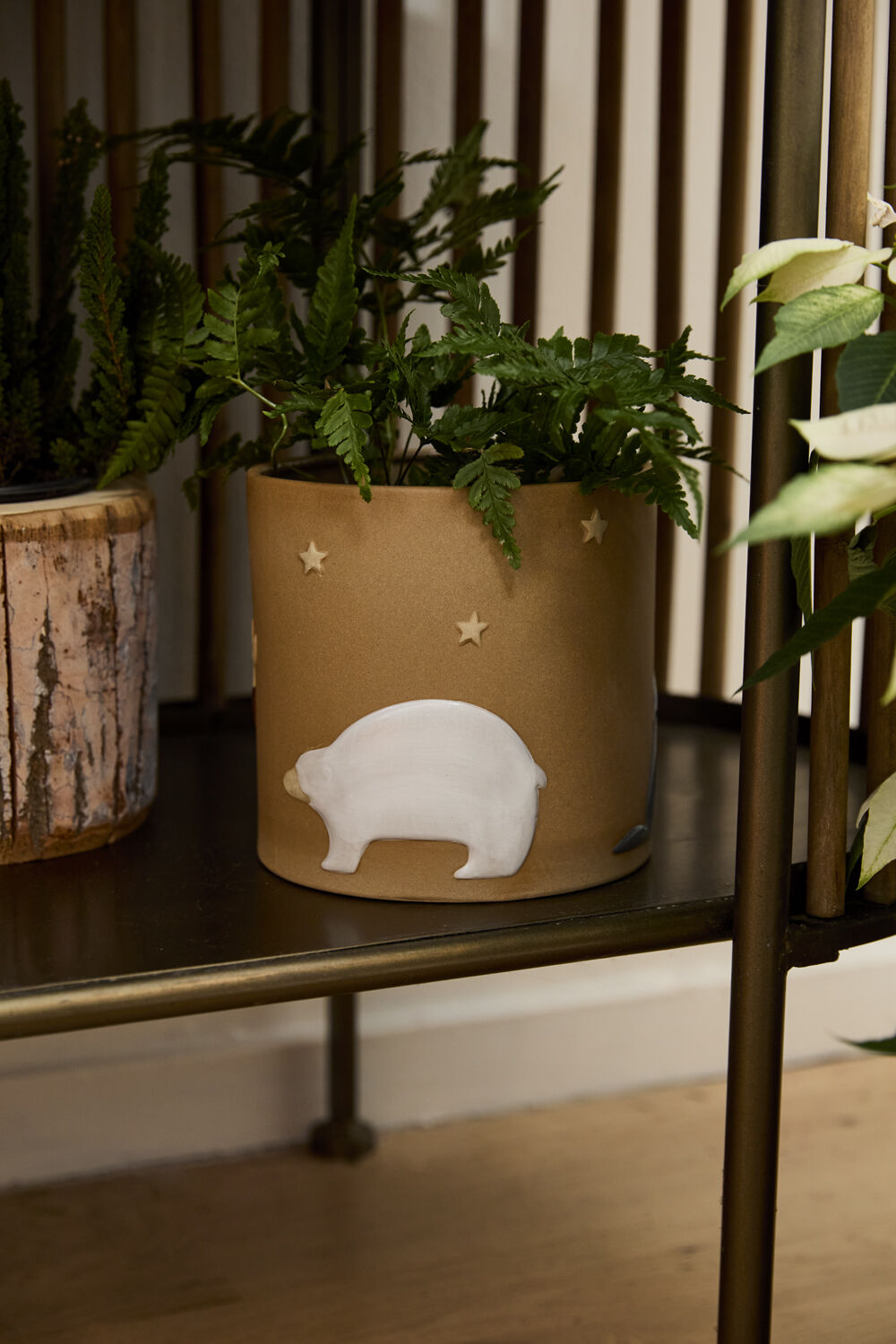 A green fern sits in the Accent Decor Antarctica Pot, a brown ceramic planter featuring a white bear and star design. The pot rests on a metal shelf beside another plant in a rustic container.