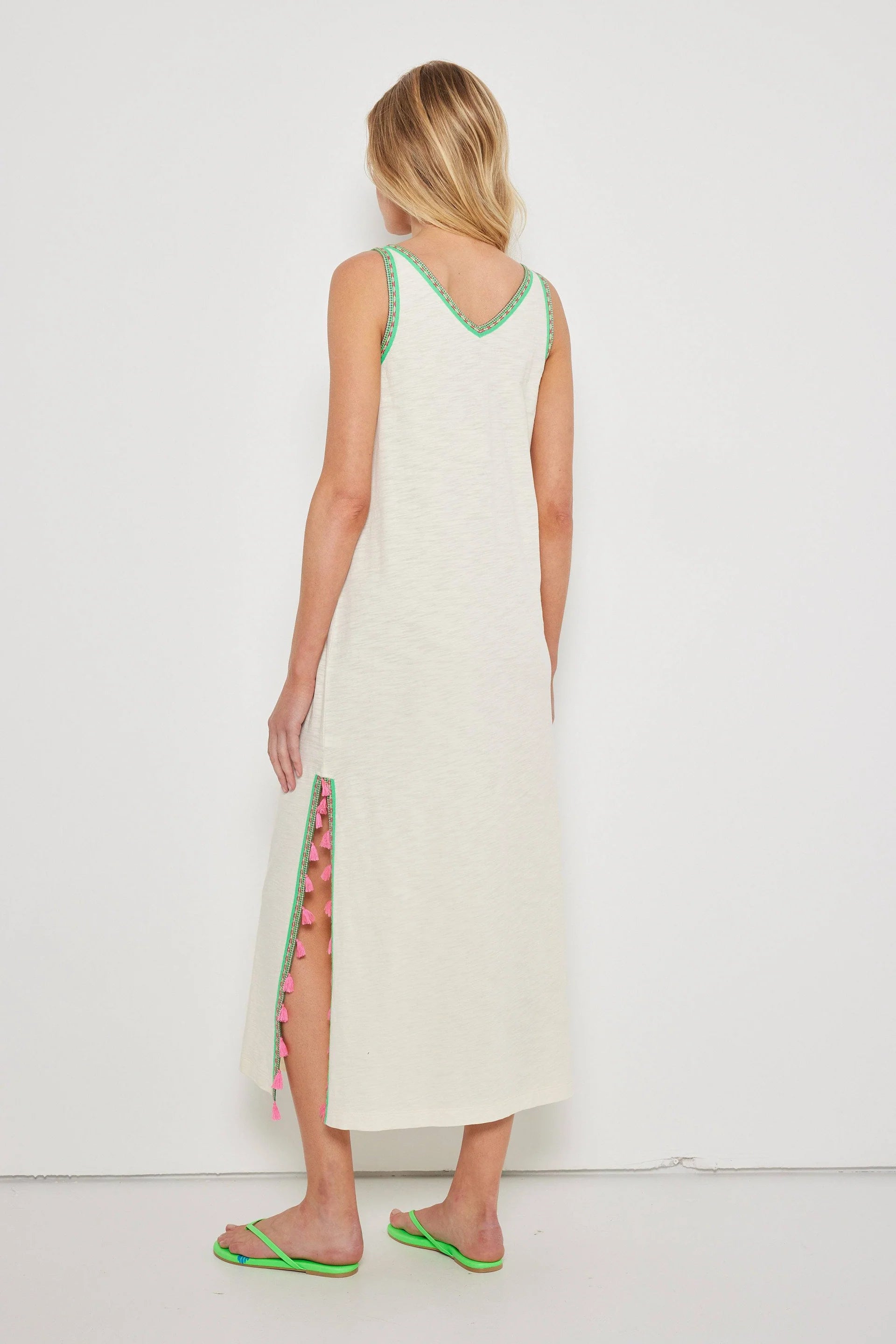 A woman with blonde hair faces away, wearing the Lisa Todd Tassel Toss Dress—a sleeveless cotton maxi with embroidered trims and pink tassels on the side slit. She pairs it with green flip-flops against a plain white background.