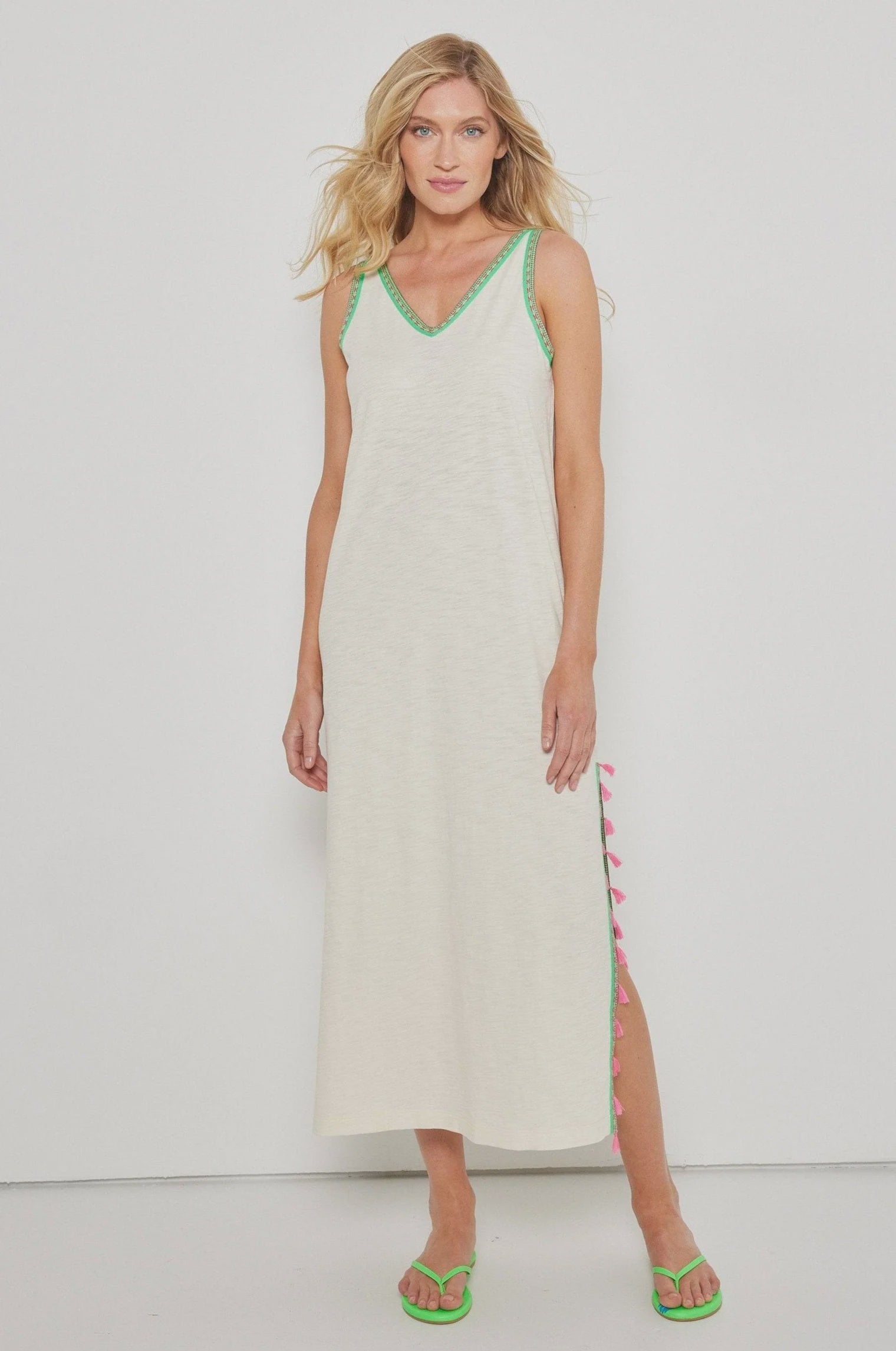 A woman stands against a white background wearing the Lisa Todd Tassel Toss Dress, a sleeveless cream maxi with green embroidered trims and pink tassels along the side, paired with green open-toe sandals.