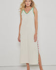 A woman stands against a white background wearing the Lisa Todd Tassel Toss Dress, a sleeveless cream maxi with green embroidered trims and pink tassels along the side, paired with green open-toe sandals.