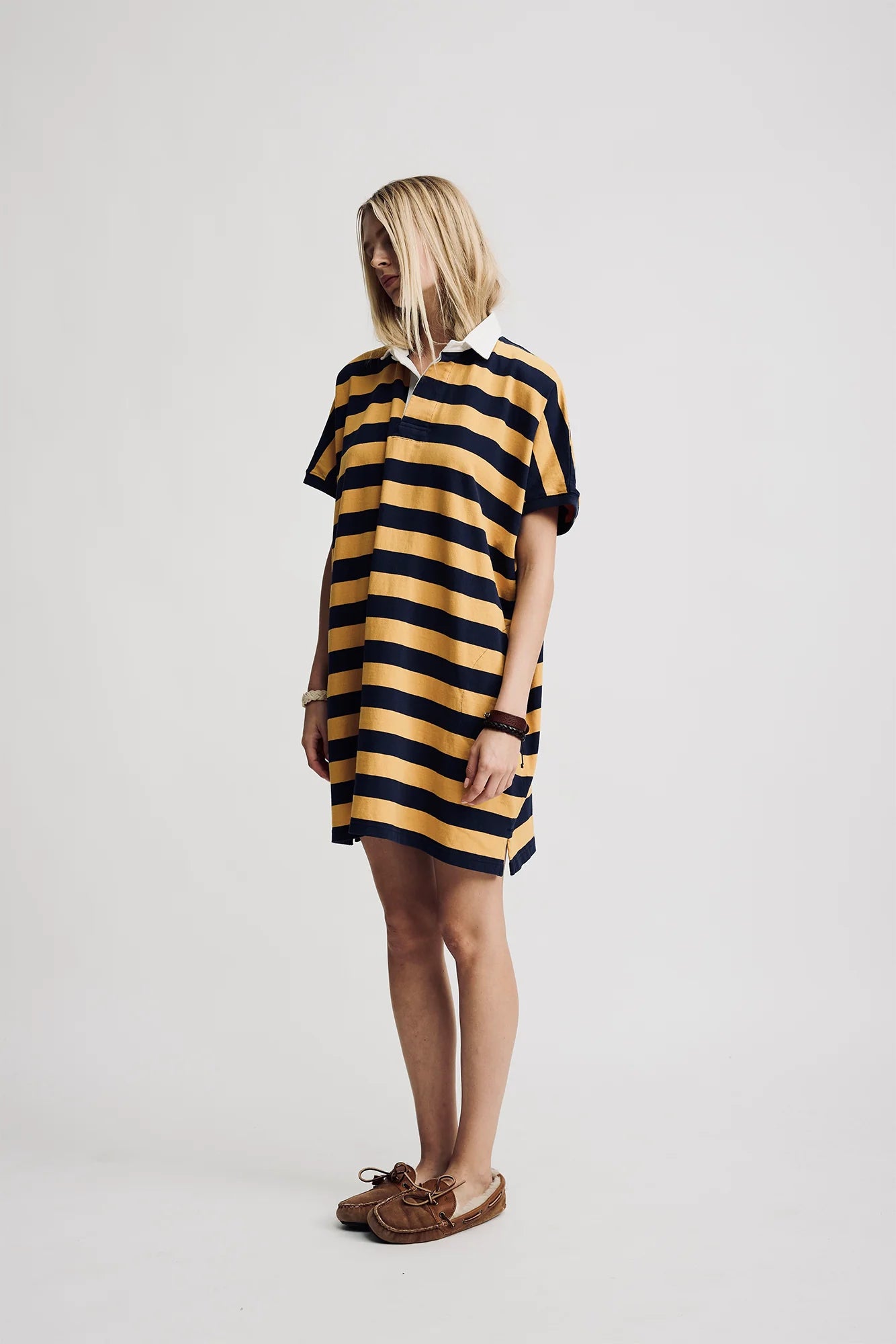 A blonde model wears the Denimist Rugby Shirt Dress—a yellow and black striped, oversized cotton piece—paired with moccasin-style shoes, standing against a plain white background.