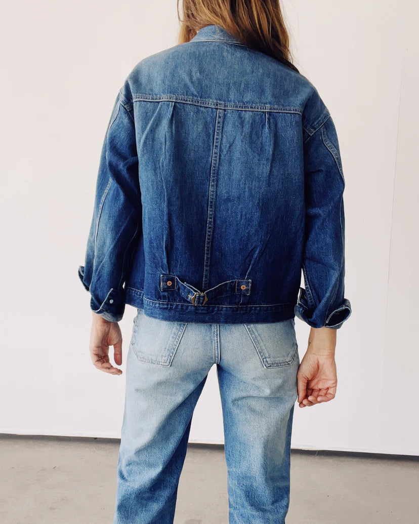 A person with long hair stands facing a white wall, wearing The Denim Jacket by Hey Gang, along with light blue jeans. The sleeves of the vintage jacket are rolled up.