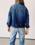 A person with long hair stands facing a white wall, wearing The Denim Jacket by Hey Gang, along with light blue jeans. The sleeves of the vintage jacket are rolled up.