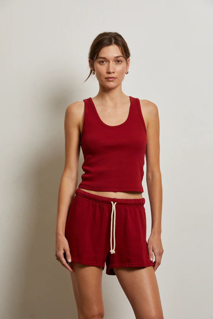 A woman with light brown hair tied back stands against a plain wall, wearing the Perfectwhitetee Layla Terry Short and a sleeveless red tank top. The shorts feature french terry cotton, an elastic waistband, and a white drawstring. She looks neutral.