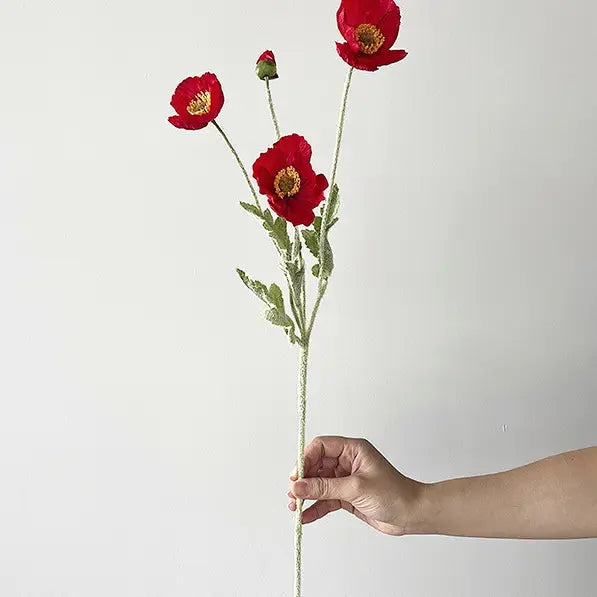 A hand holds the Faire 22'' Single Flocked Floral Poppy Spray, displaying three vibrant red flowers and one bud on a slender stem against a plain white background.
