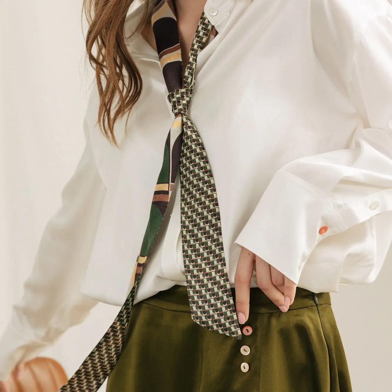 A person in a white button-up shirt with long sleeves and an abstract patterned tie epitomizes high fashion. They pair this with olive green pants adorned with colorful buttons, while one hand skillfully adjusts their Modular Silk Skinny Scarf in green from Faire.