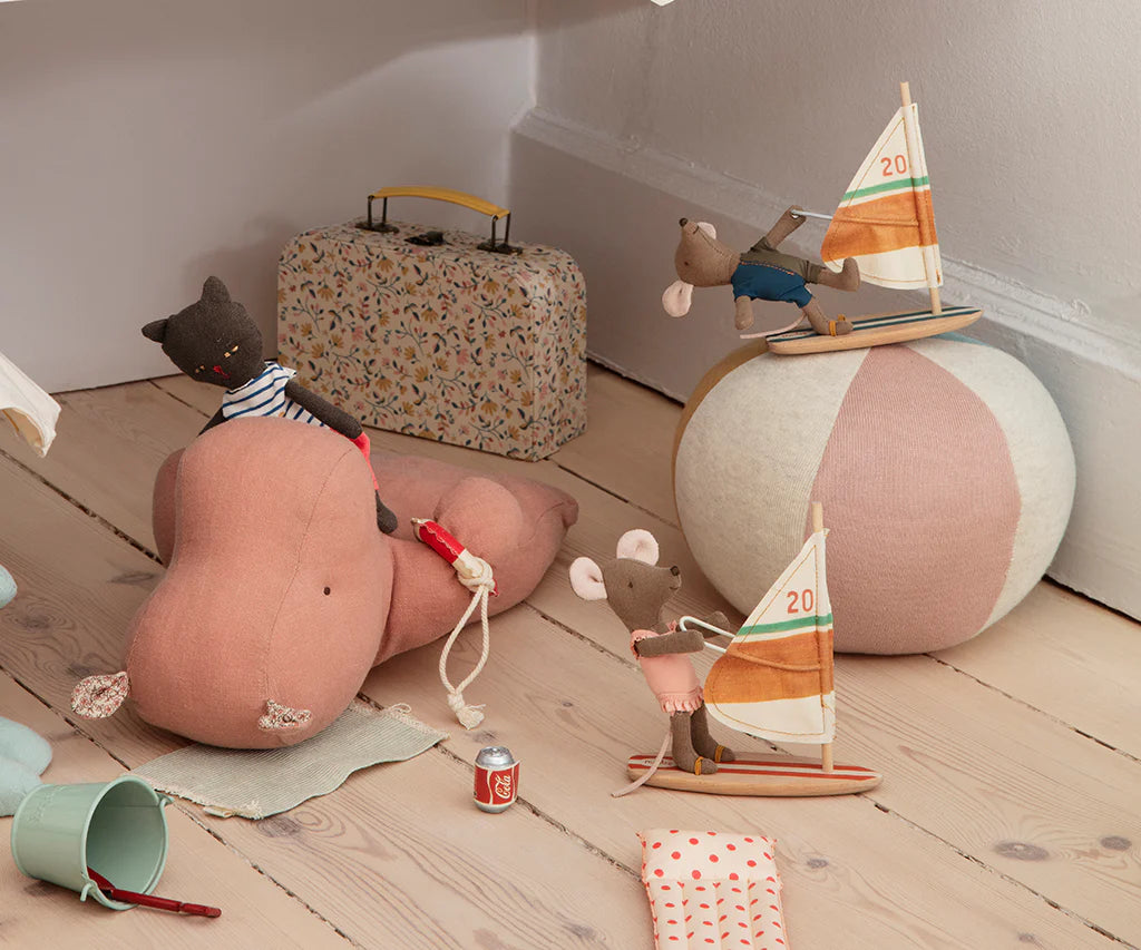 Maileg stuffed animals, like mice with sailboats and the Maileg Little Sister/Beach Surfer, are playfully arranged on a wooden floor with a pastel beach ball, patterned suitcase, cans, fabric pieces, and a cat riding a hippo.