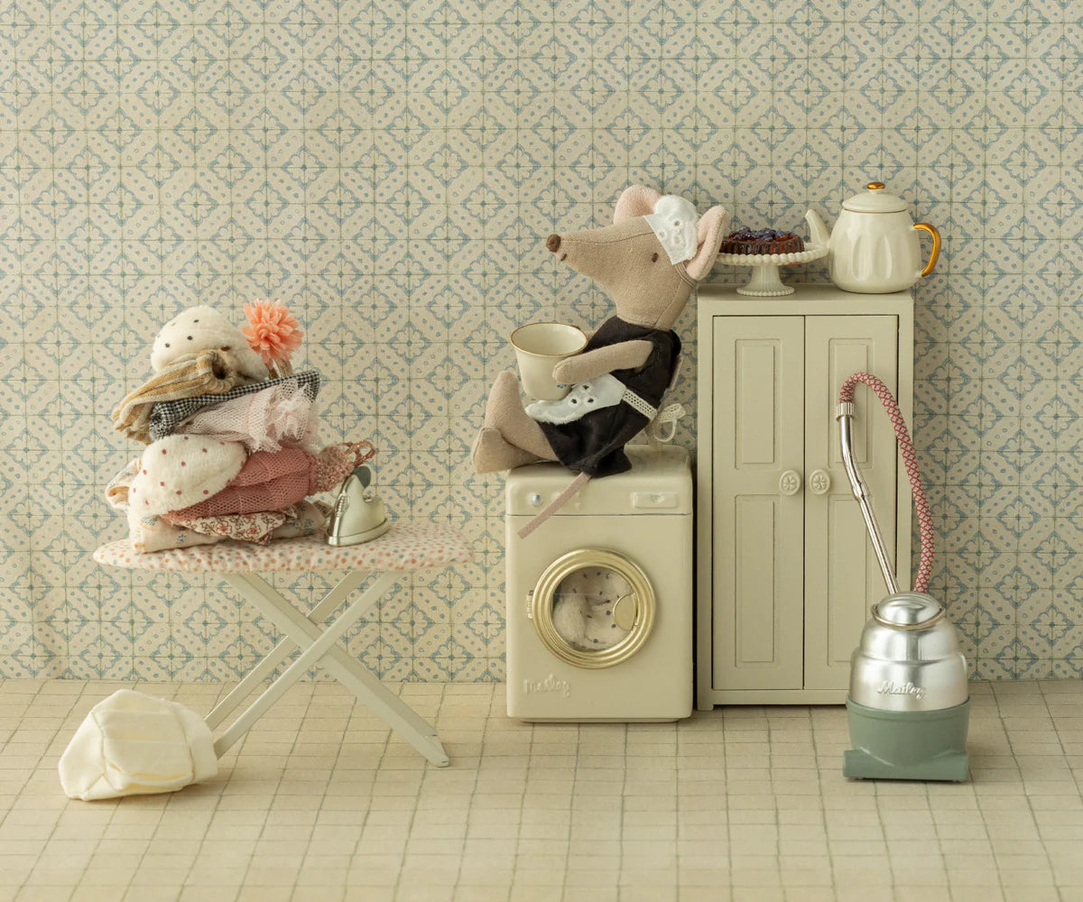 A whimsical Maileg scene: the Maileg Maid Mouse / Big Sister sits on a washing machine, another mouse holds a laundry basket on an ironing board, surrounded by props like an iron and teapot, all set against a charming patterned background.