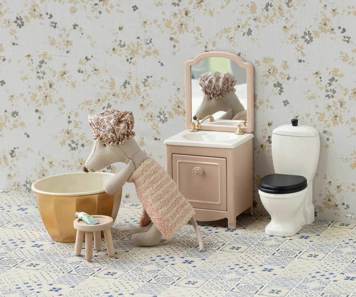 The Maileg Wellness Mouse / Big Sister by Maileg stands in a cute playset bathroom with a sink, mirror, toilet, and stool holding a toothbrush. She wears a towel and shower cap as she leans over the bowl on the patterned floor.