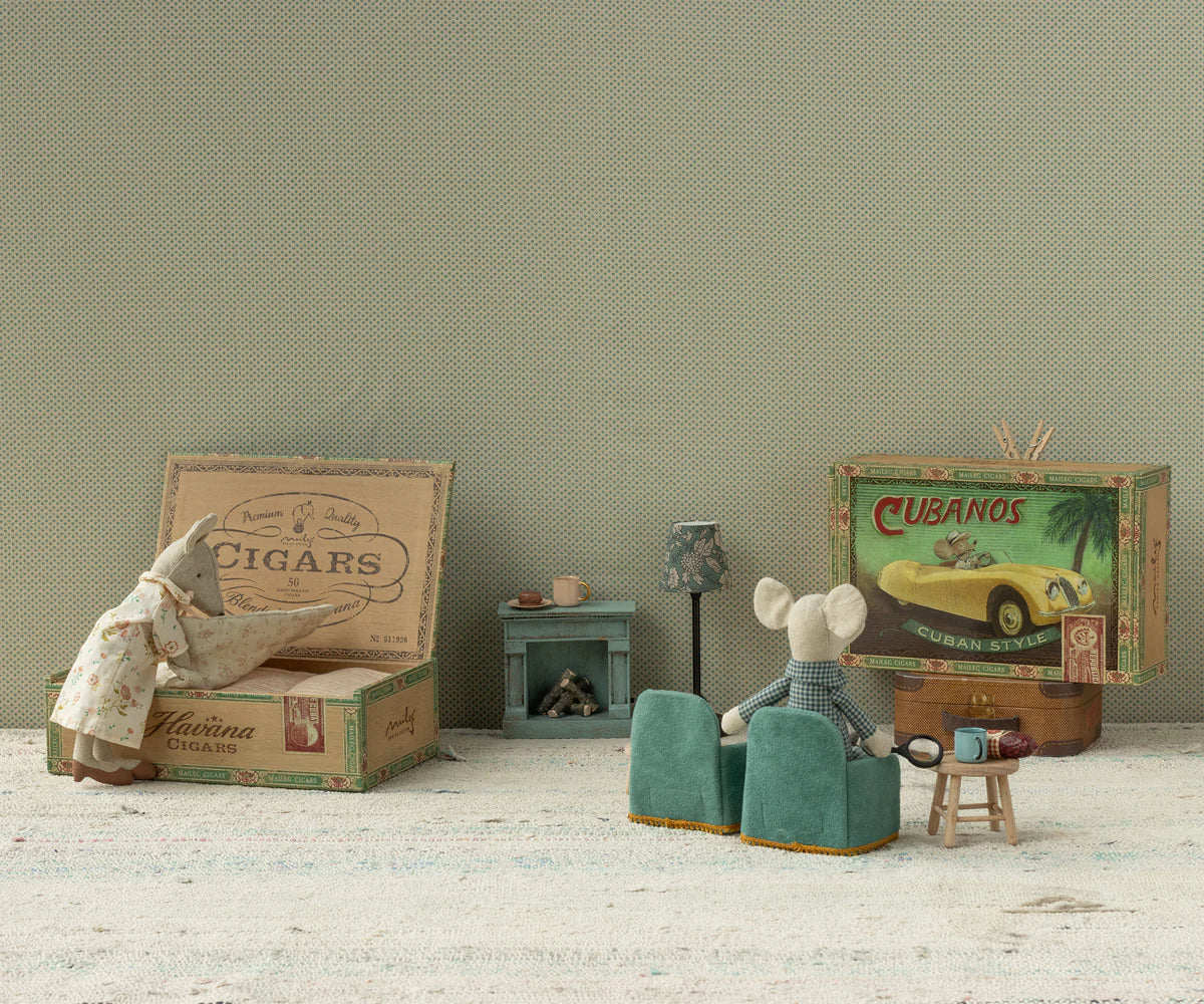 Maileg Grandma & Grandpa Mice by Maileg relax in a miniature living room with tiny chairs, a table, lamp, fireplace, and even a matchbox bed—one opens a box while the other lounges in an armchair.