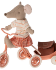 The Maileg Tricycle Mouse, Big Sister by Maileg rides a pink tricycle with white wheels and a matching cart. Her striped shirt and plaid skirt fasten with hidden magnets, and her small backpack rests in the cart.