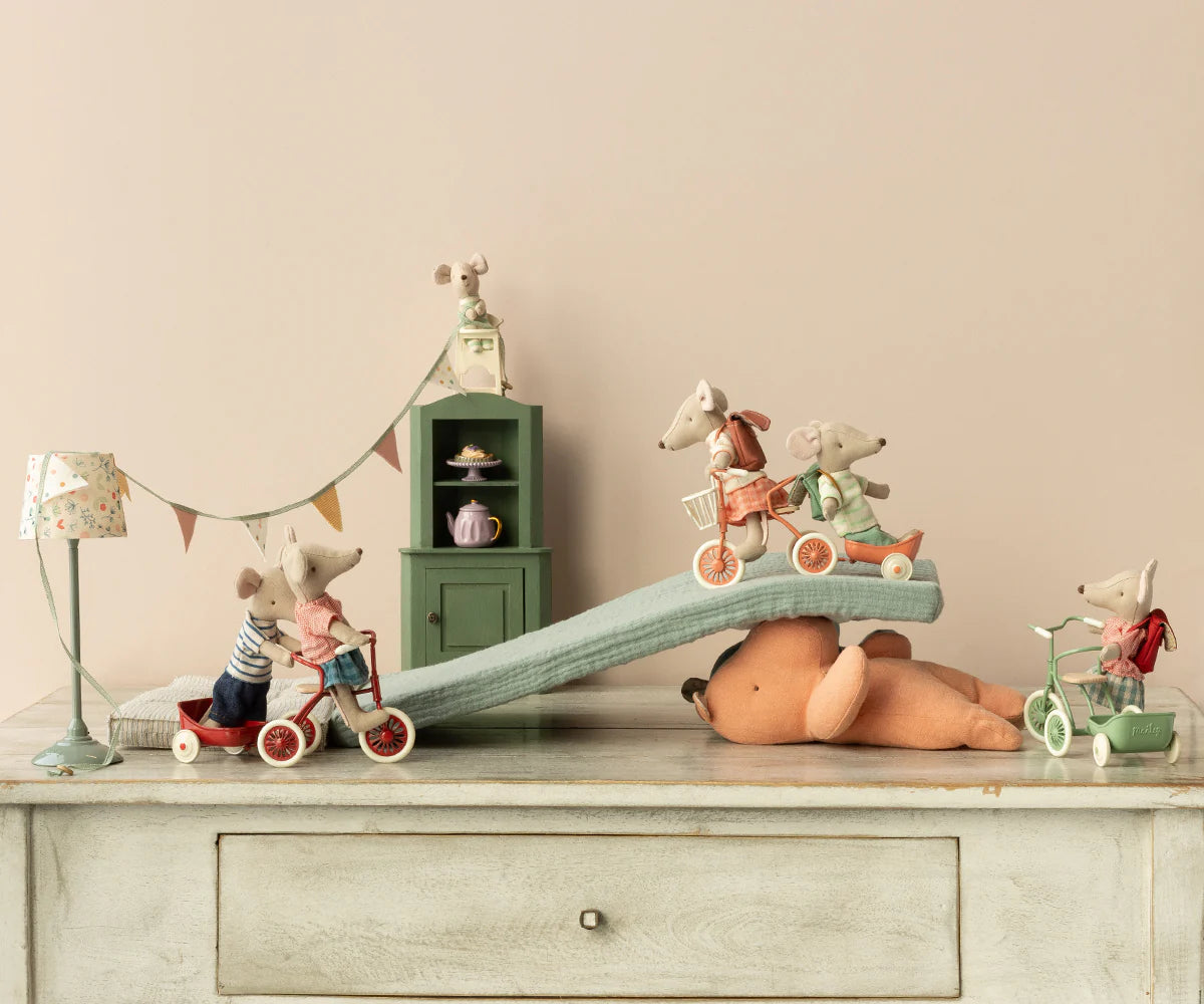 Five Maileg toy mice, including the Maileg Tricycle Mouse, Big Sister, ride bikes and tricycles down a cushion ramp on a plush bear, with magnets securing the scene atop a vintage-style dresser near a dollhouse cabinet and lamp.