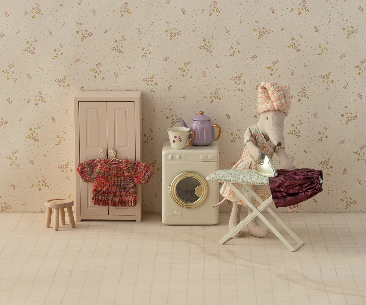 The Maileg Skirt &amp; Knitted Blouse mouse by Maileg, dressed in a skirt and blouse, irons clothes on a dollhouse ironing board next to a washing machine, cupboard, stool, and teapot in a charming laundry room scene.