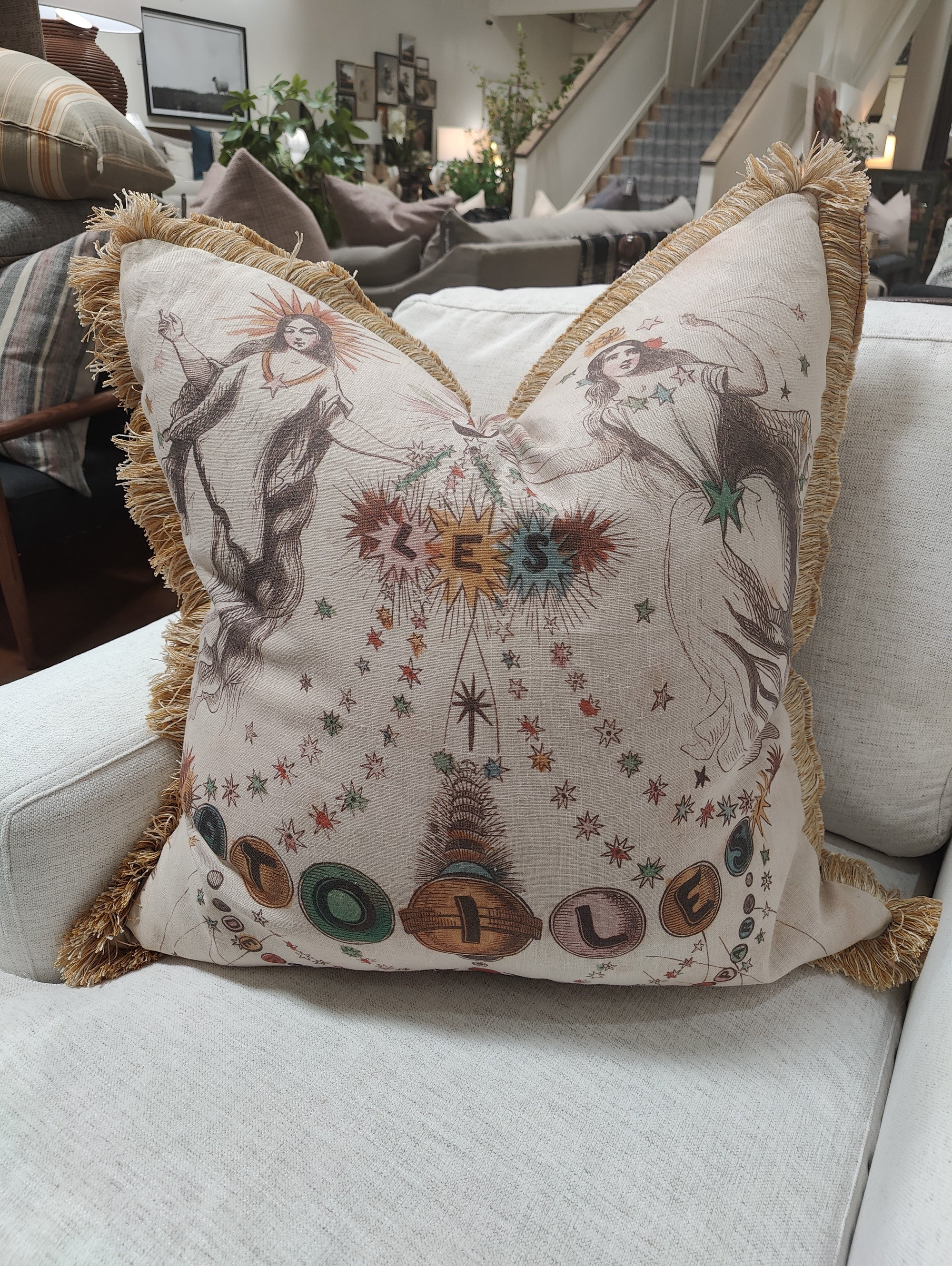 The Design Legacy Les Etoiles Maize Fringe 26x26 pillow features two women surrounded by stars, "Bless Home" in colorful letters, and makes a decorative accent on a white couch.
