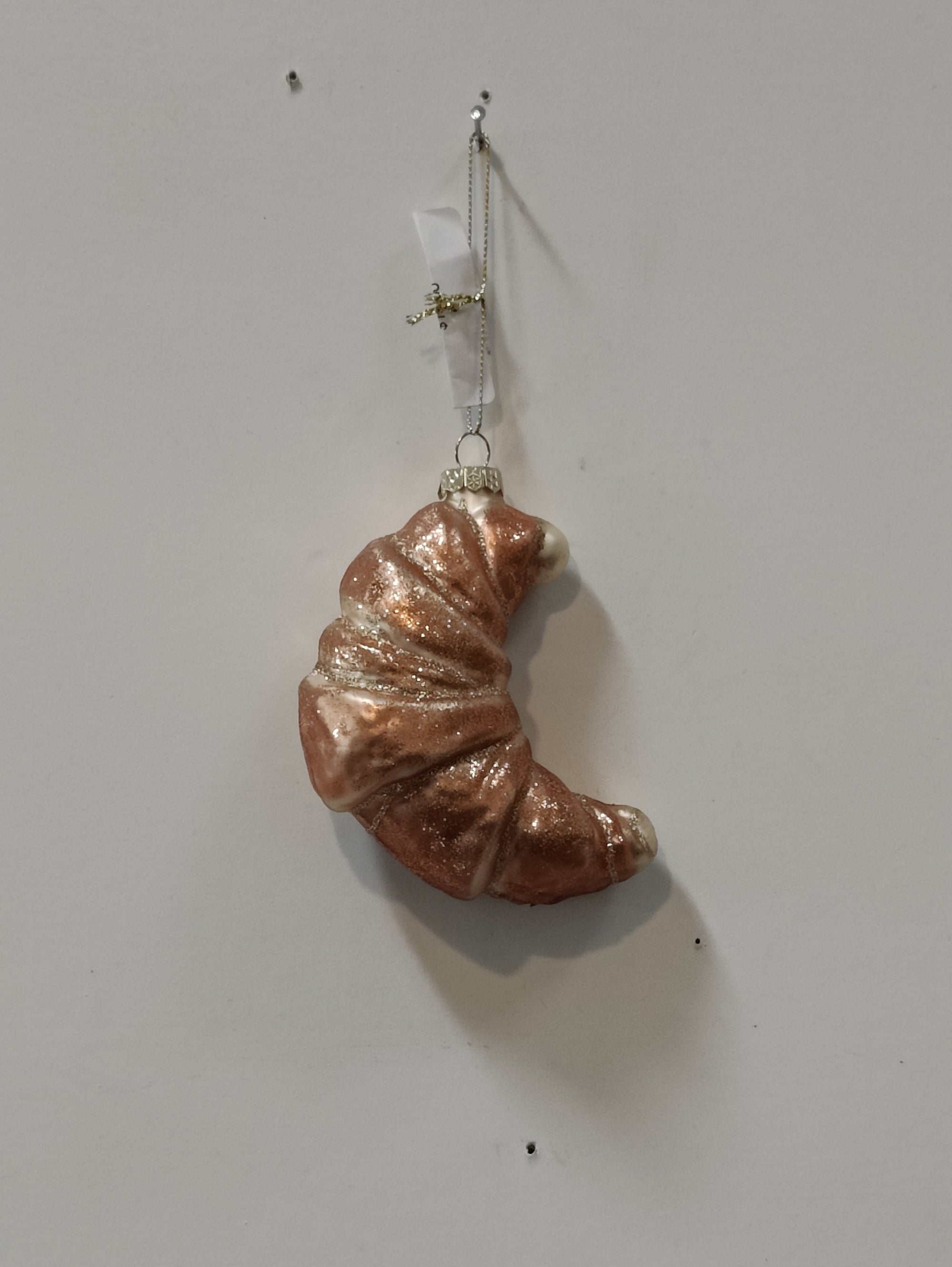 The Croissant Ornament by Creative Co-op, with its shimmering metallic finish, hangs against a plain white wall. It features a glittery croissant shape and comes complete with a small hook and loop for easy hanging.