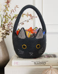 The Accent Decor Tricks & Treats Basket, shaped like a black cat with yellow eyes and gray ears and filled with colorful candies, sits on stacked books beside a vase of flowers and a decorative pumpkin for festive Halloween decor.